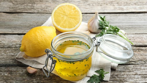 Glass jar of homemade dressing with herbs and oil on a rustic wooden table, surrounded by a whole lemon, a halved lemon, a garlic bulb, parsley, and a small cloth napkin.