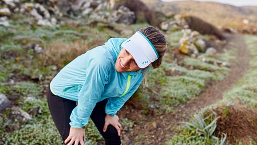 A woman stretching while bending forward on a narrow outdoor trail, wearing a light blue long-sleeve athletic top.