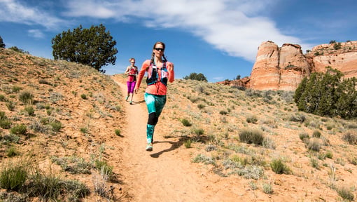 Two trail runners moving downhill on a narrow desert path, with one in the foreground wearing a hydration vest and sunglasses.