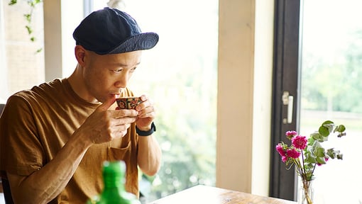 Man drinking herbal tea from a cup at a table with flowers near a window.