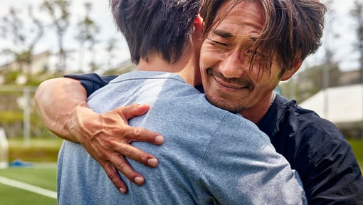 Two friends hugging outdoors with greenery in the background.