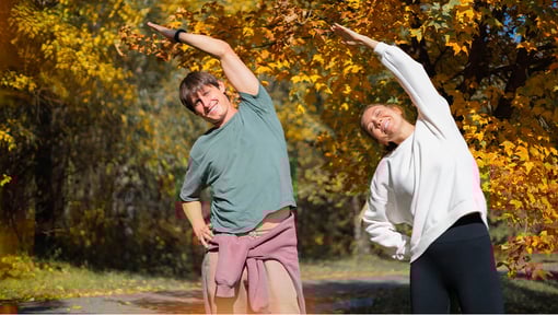 Two people doing side stretches outdoors among fall foliage.