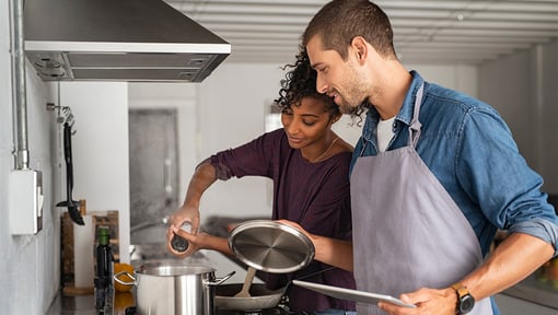 Two people cooking chicken soup together in a modern kitchen, one stirring a pot on the stove while the other holds a lid and a tablet.