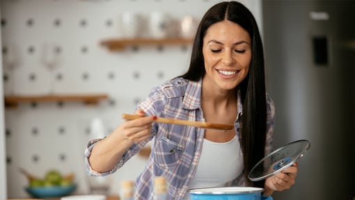 A woman in a kitchen holding a pot lid and tasting chicken quinoa soup from a wooden spoon over a blue cooking pot