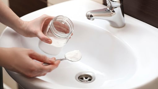 Hands holding a glass jar and a spoon with baking soda over a bathroom sink, preparing to use it for cleaning.