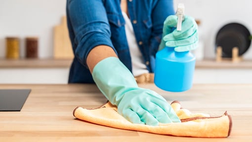 Person wearing green rubber gloves cleaning a wooden surface with a yellow cloth, holding a blue spray bottle filled with DIY all-purpose cleaner in the other hand.