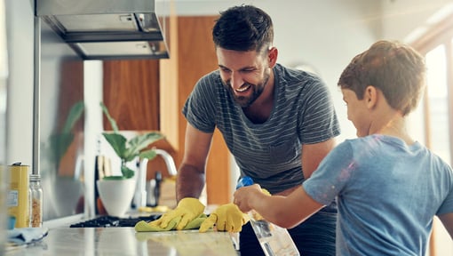 A man wearing yellow cleaning gloves scrubbing a kitchen counter while his child stands nearby, spraying a natural cleaning solution on the countertop.