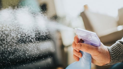 Close-up of a hand spraying liquid from a blue spray bottle, with mist dispersing into the air, for cleaning purposes.
