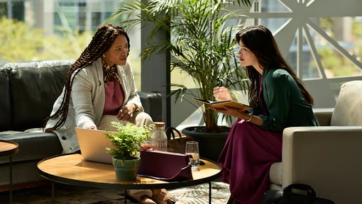Two coworkers sitting in a modern lounge area with natural light, chatting.