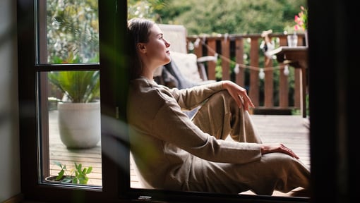 Person sitting in a doorway with legs stretched out, leaning back and gazing toward a wooden deck surrounded by greenery, suggesting a calm and relaxing moment.