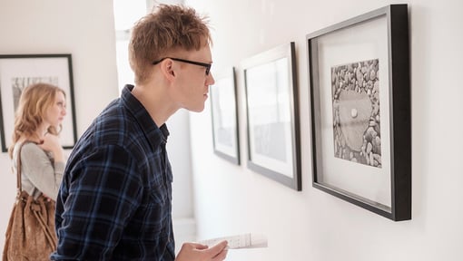 Person closely examining framed artwork on a white wall inside an art gallery with another person viewing art in the background.