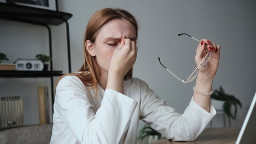 A woman sitting at a desk holding eyeglasses in one hand and resting the other hand on their head, appearing overwhelmed while working on a laptop, representing burnout.