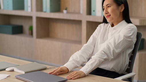 Woman seated at a desk in an office setting with hands resting near a closed laptop, wearing a white long-sleeve shirt.