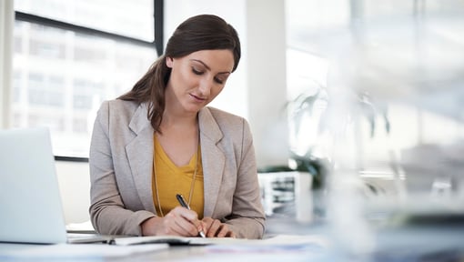Woman sitting at a desk in a bright office environment, writing on paper next to an open laptop.