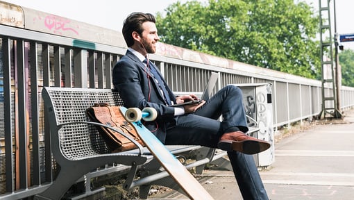 Man in a business suit sitting on a bench at an outdoor train station platform, working on a laptop with a longboard skateboard resting against the bench.