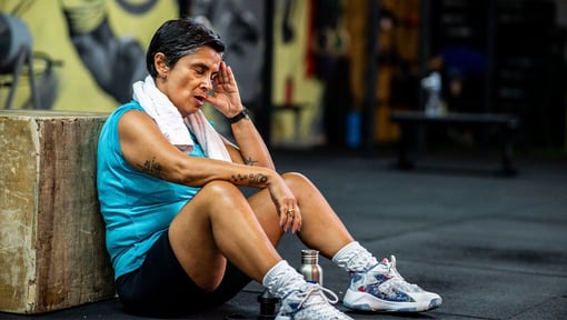 Person resting on gym floor after workout, sitting against a wooden box with towel and water bottle nearby.