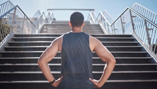 Man in athletic gear standing at the bottom of an outdoor staircase, looking upward with hands on hips, preparing for a workout.