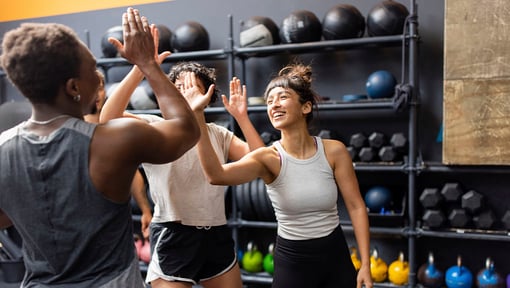 Group of people in athletic wear giving high-fives in a gym, with kettlebells and medicine balls arranged on shelves in the background.