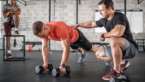 A fitness trainer assisting a man performing a dumbbell plank row exercise in a gym.