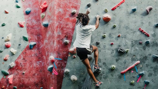Person climbing an indoor rock wall with colorful handholds and footholds, wearing athletic shorts and a t-shirt.