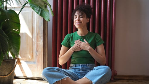 Woman sitting cross-legged indoors with hands placed over the chest in a gratitude practice.