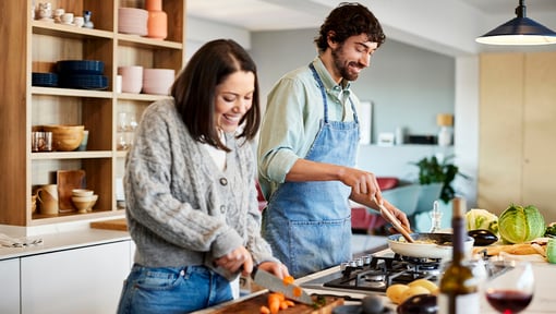 Two people cooking together in a modern kitchen; one is chopping vegetables on a cutting board while the other is stirring food in a pan on the stove.