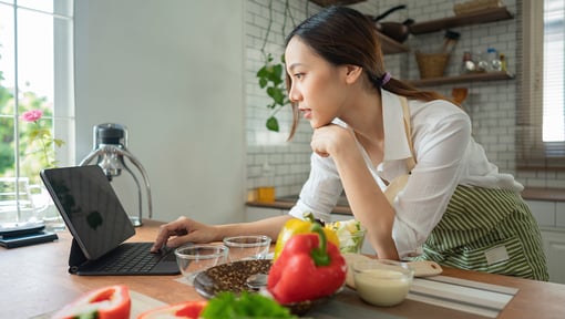A woman wearing an apron in a kitchen, looking at a tablet preparing to make a recipe with fresh vegetables and ingredients.