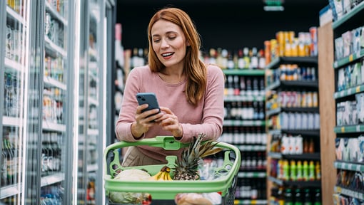 A woman in a grocery store aisle pushing a green shopping cart filled with fresh produce, holding a smartphone.