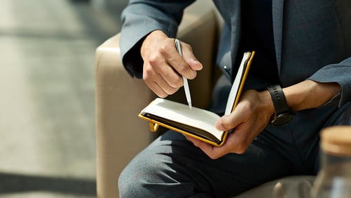 Person dressed in a dark suit sitting on a chair, writing in an open notebook with a pen.