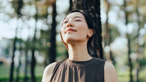 A woman standing outdoors among tall trees. Her head is slightly tilted upward, suggesting a serene moment.