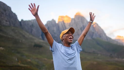 Person standing outdoors in front of a mountain range, wearing a light gray T-shirt and a yellow cap, with both arms raised high in the air. 