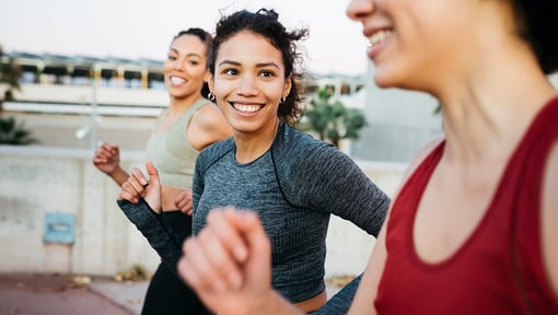 Three women jogging outdoors on a paved path, the women are smiling.