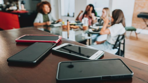 Several smartphones placed on a wooden surface in the foreground, with a group of people sitting around a dining table in the background. 