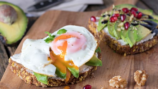 Two slices of multigrain bread topped with avocado. One slice has a sunny-side-up egg with a runny yolk, and the other slice is garnished with avocado slices, pomegranate seeds, and seeds. 