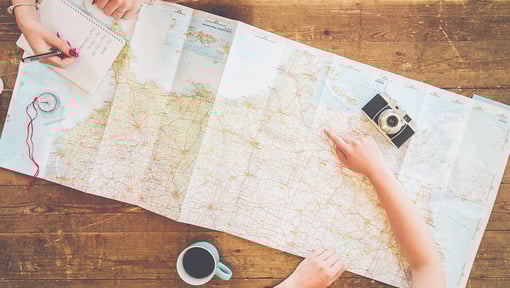 A top down view of two people sitting at a wooden table planning a trip with a large folded map. One person is writing in a notebook while the other points to a spot on the map