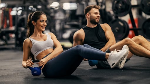 Two people exercising on the gym floor with kettlebells, illustrating an active lifestyle supported by eating foods that promote health and fitness. 