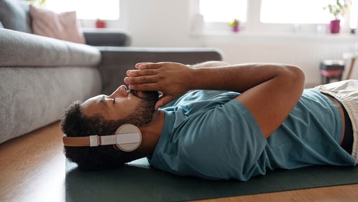 A man lying on a yoga mat at home wearing headphones, practicing relaxation and mindful rest.
