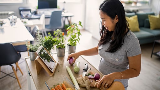 A woman preparing vegetables at a kitchen counter while following a recipe on a tablet, with fresh herbs and ingredients nearby.