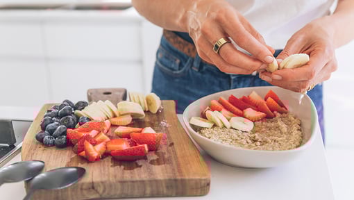 Hands preparing a bowl of oatmeal topped with sliced strawberries and bananas beside a cutting board with fresh fruit.