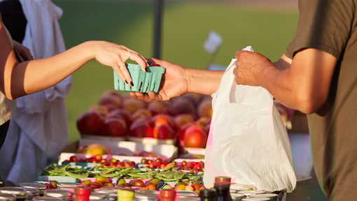 Hands exchanging a small container of berries at an outdoor farmers market stand with fresh produce displayed on a table.