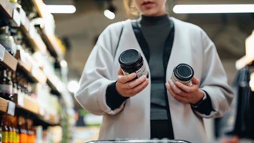 Person standing in a grocery store aisle comparing two packaged food products while shopping.