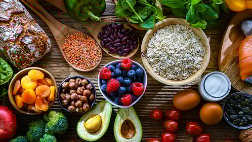 Assortment of whole foods including fruits, vegetables, grains, nuts, and bread arranged on a wooden table.