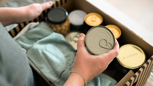 Hands place canned food and folded clothing into a donation box, preparing supplies for a community food or clothing drive.