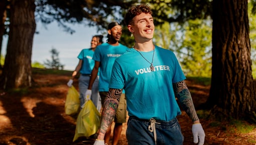 A group of volunteers wearing matching blue volunteer shirts walk along a wooded trail during an outdoor cleanup activity.