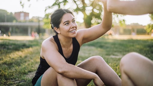 A woman is sitting in the grass after a workout high-fiving someone out of frame.