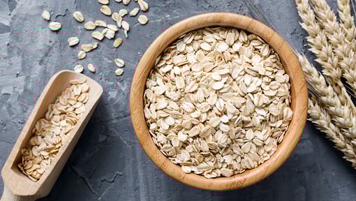 Overhead view of whole grain oats in a wooden bow, with a scoop of oats and wheat stalks nearby.