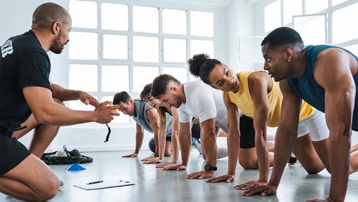 Fitness instructor timing and coaching a group of adults performing plank exercises in a gym.