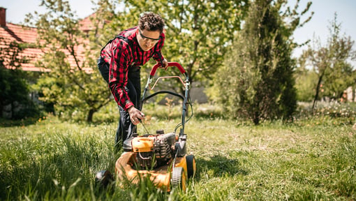 A man mowing an overgrown lawn with a push mower in a residential outdoor setting.