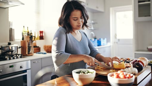 A woman preparing a meal in a bright home kitchen, chopping ingredients on a cutting board with bowls of fresh produce on the counter.