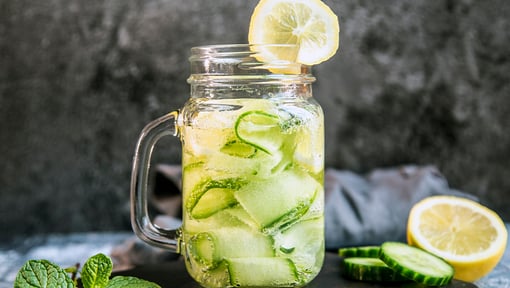 Glass mason jar filled with cucumber and lemon–infused water, garnished with a lemon slice, set against a dark background with fresh mint.